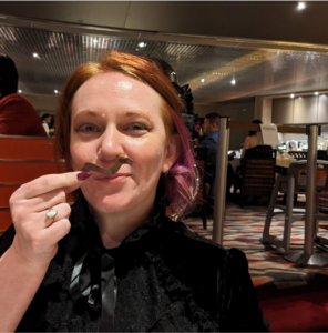 Photo of Leslie holding a fake mustache (made of chocolate) in front of her face, smiling at the silliness of it. She has short red hair with red-violet accents and is wearing a high collared black gown of lace and velvet.